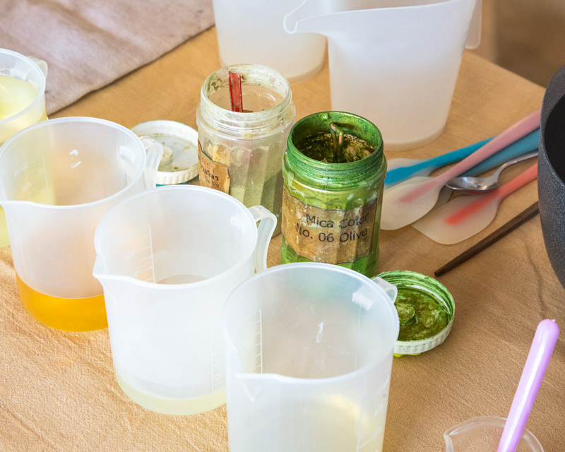 Soap-Making Accessories and Ingredients | Sabai Home A collection of soap-making tools and ingredients, including plastic beakers, mica oil, a spoon, and other essentials.