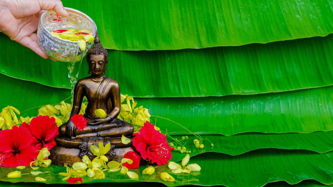 A hand pouring scented water with flower petals over a Buddha statue as part of a traditional Songkran blessing ritual in Thailand.