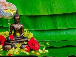 A hand pouring scented water with flower petals over a Buddha statue as part of a traditional Songkran blessing ritual in Thailand.
